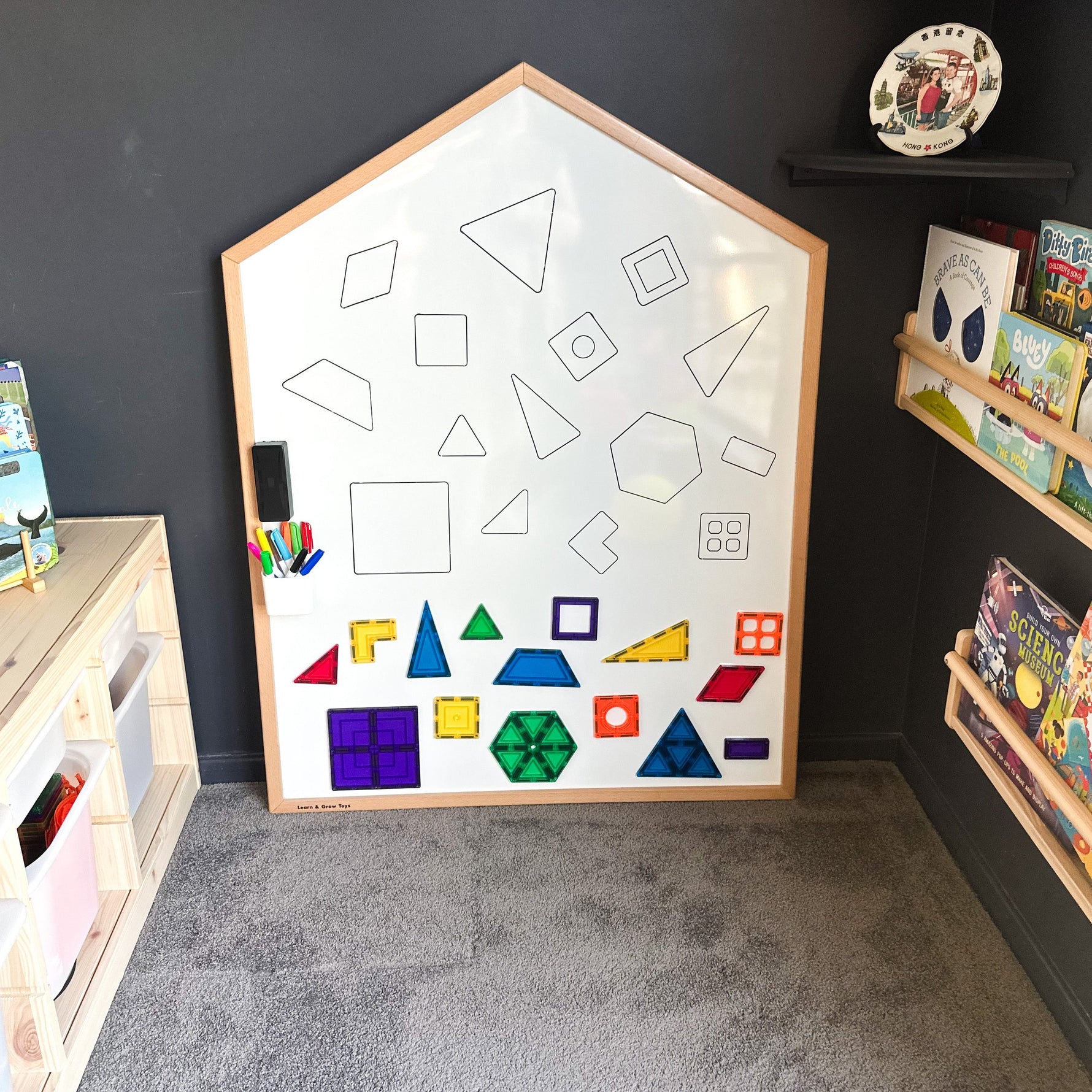 Learn & Grow Toys Multi-Board with shapes drawn on whiteboard sitting in a dark playroom with books displayed on wall