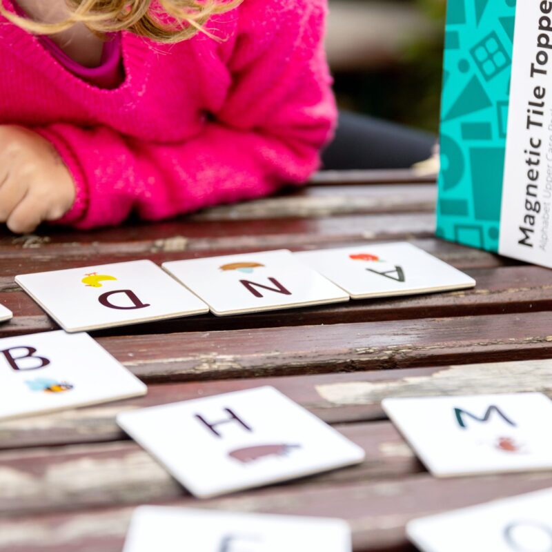 Child in pink jumper playing with alphabet uppercase tile toppers on a wooden table outside