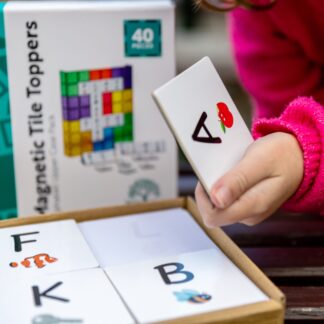 Child in pink jumper holding the A from the alphabet uppercase tile toppers pack on a wooden table outside