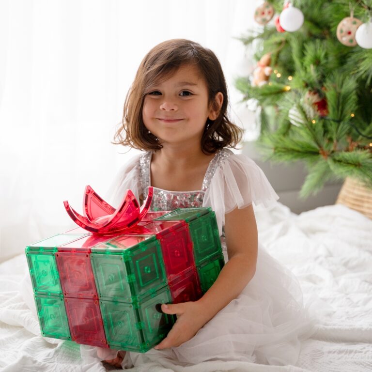 Child dressed in white holding christmas gift made from green and red magnetic tiles from learn and grow toys with christmas tree in background