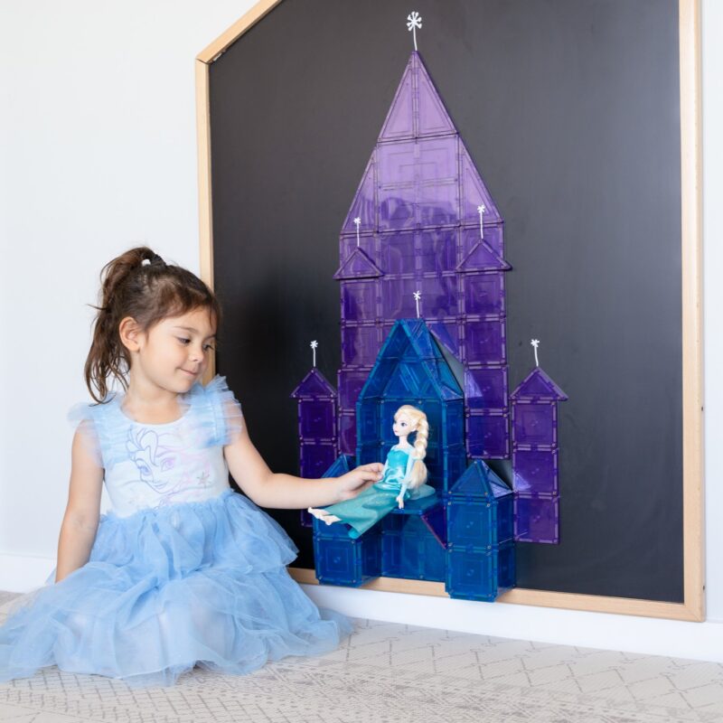 Child in Frozen Elsa costume holding Elsa doll against chalkboard of Multi-Board against the wall